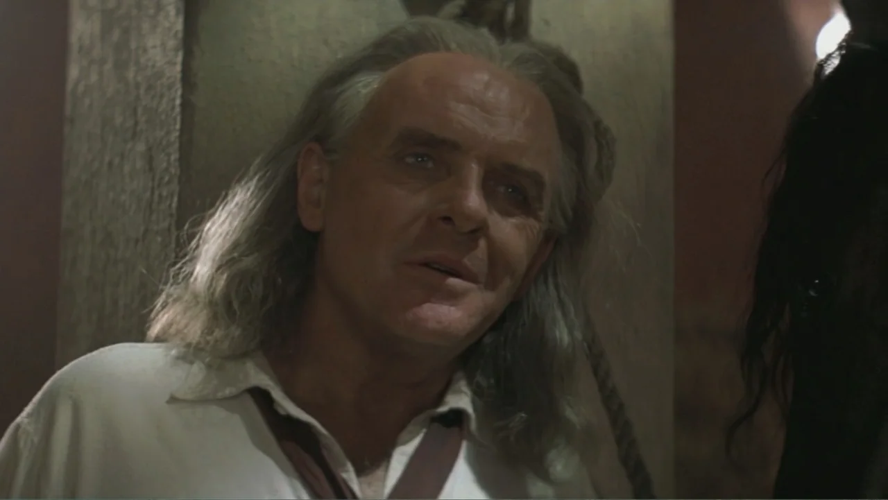 The Mask of Zorro (1998) movie still: closeup — A dark stone cell with a rope hanging against a pillar in the background. An older man with long…