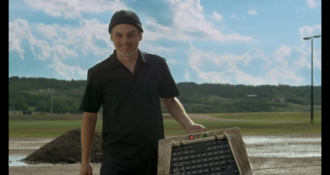 A.R.C.H.I.E. (2016) movie still: medium — A man stands in an open field under a cloudy blue sky. He holds a white plastic animal crate at his side…