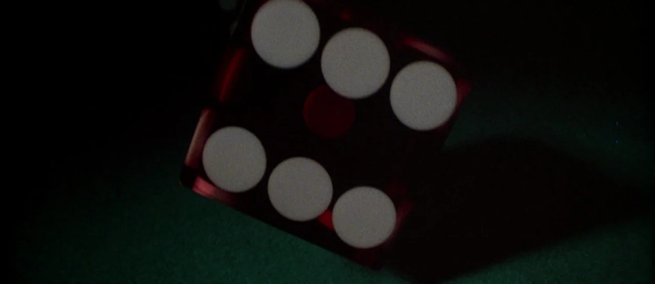 Casino (1995) movie still: closeup — A red translucent die with five white pips rests on a dark green felt surface. The object is angled slightly in…