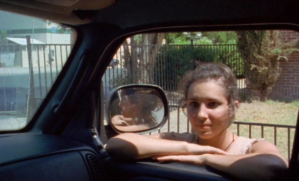 The Brown Bunny (2003) movie still: medium — A woman leans her arms against the open window of a parked vehicle. In the background a metal fence,…