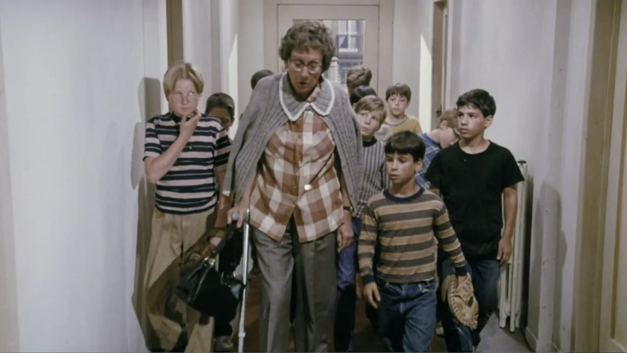 Aunt Mary (1979) movie still: fullbody — A narrow apartment building hallway features light-colored walls and a wooden door at the end. An older…