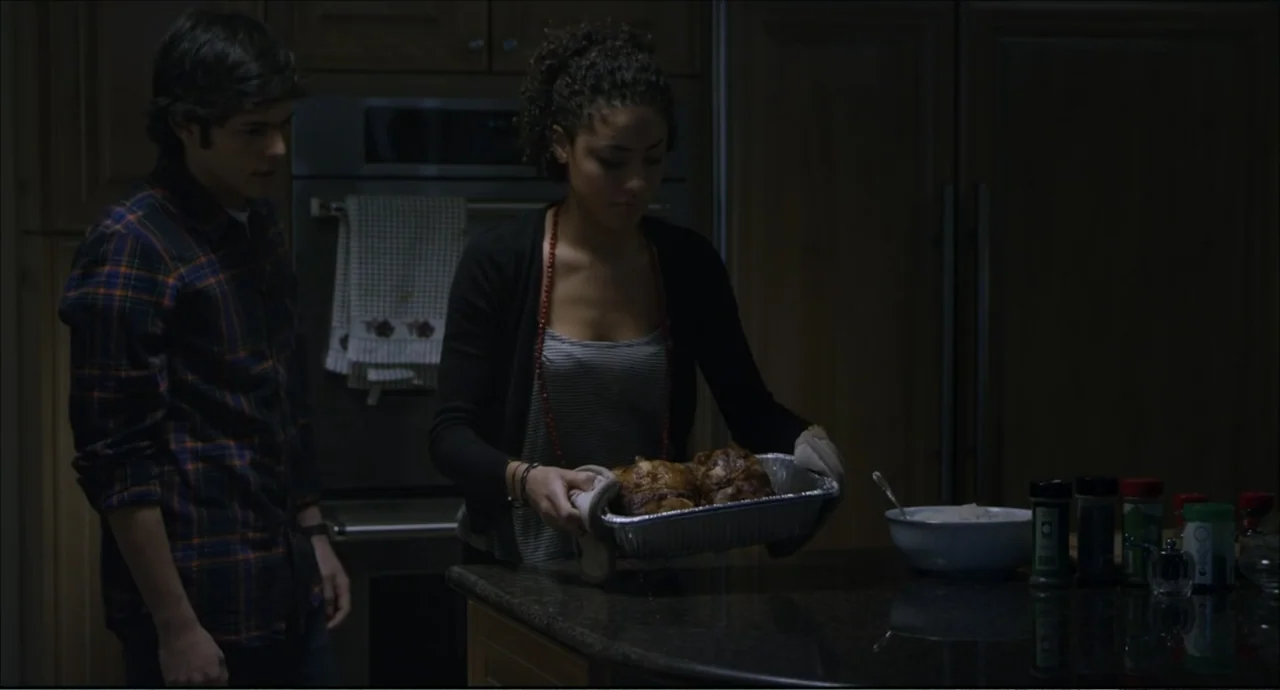 Freedom (2024) movie still: medium — A dimly lit kitchen features dark wooden cabinets and a stone countertop. A young woman places a foil roasting…