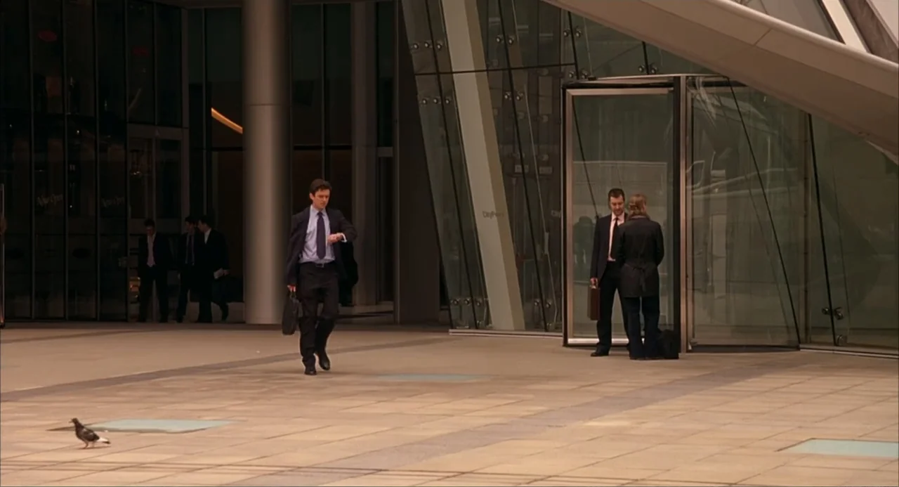 Scoop (2024) movie still: fullbody — An open stone plaza sits outside a large glass and steel corporate skyscraper. A man in a business suit walks…