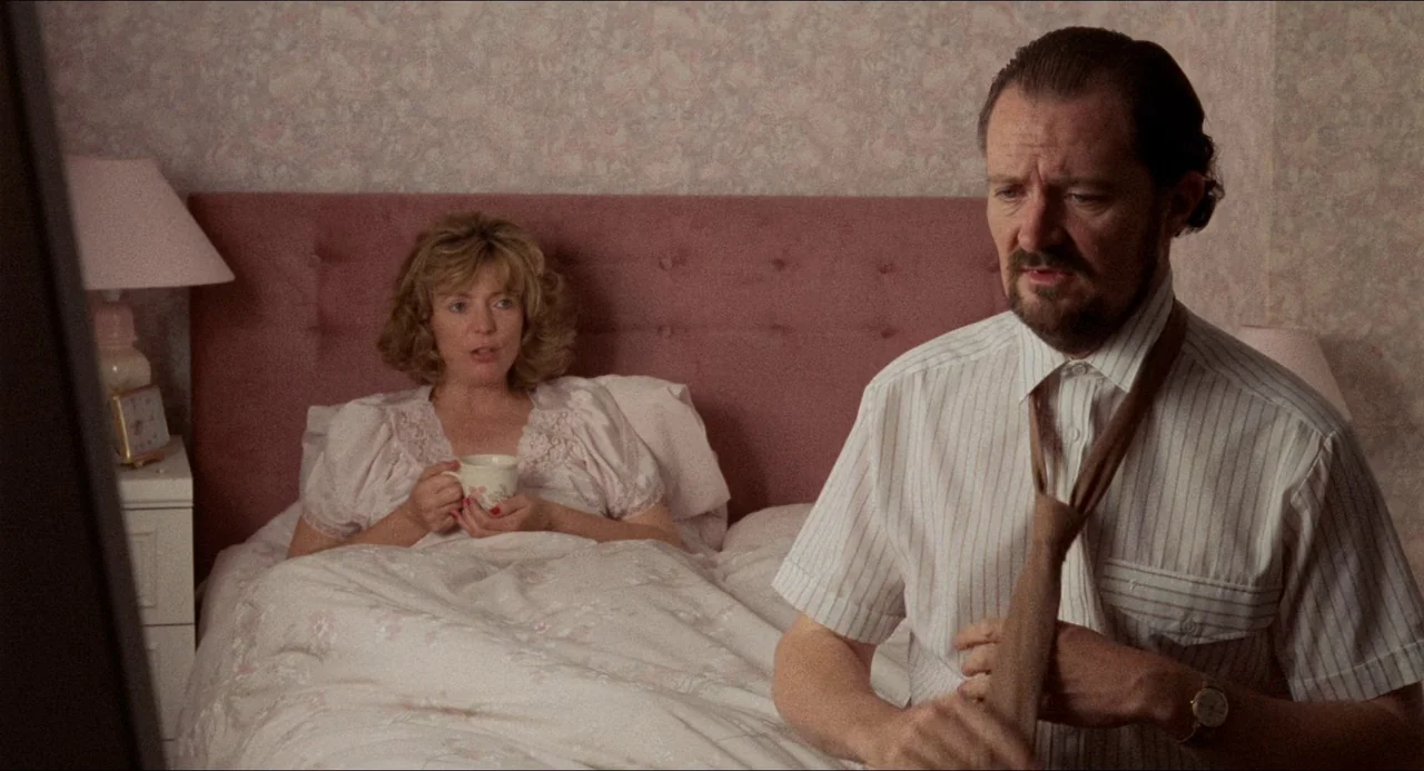 Life Is Sweet (1990) movie still: medium — A residential bedroom with floral patterned wallpaper and a large pink fabric headboard. A man stands in…