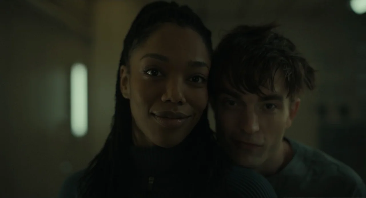 Mickey 17 (2025) movie still: closeup — A dimly lit interior room with soft shadows surrounding the two subjects. A woman and a man pose closely…