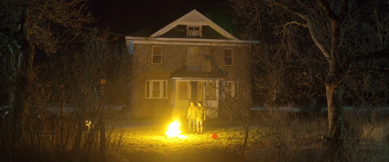 The Void (2016) movie still: wide — A two-story stone house stands behind a pair of men in a dark yard. The men observe a small, intense fire…