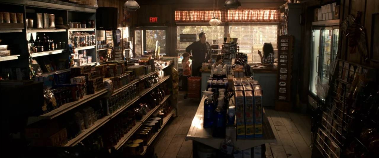 2012 (2009) movie still: wide — A rural convenience store interior with wooden walls and aisles of stocked goods. A man and a woman interact near…