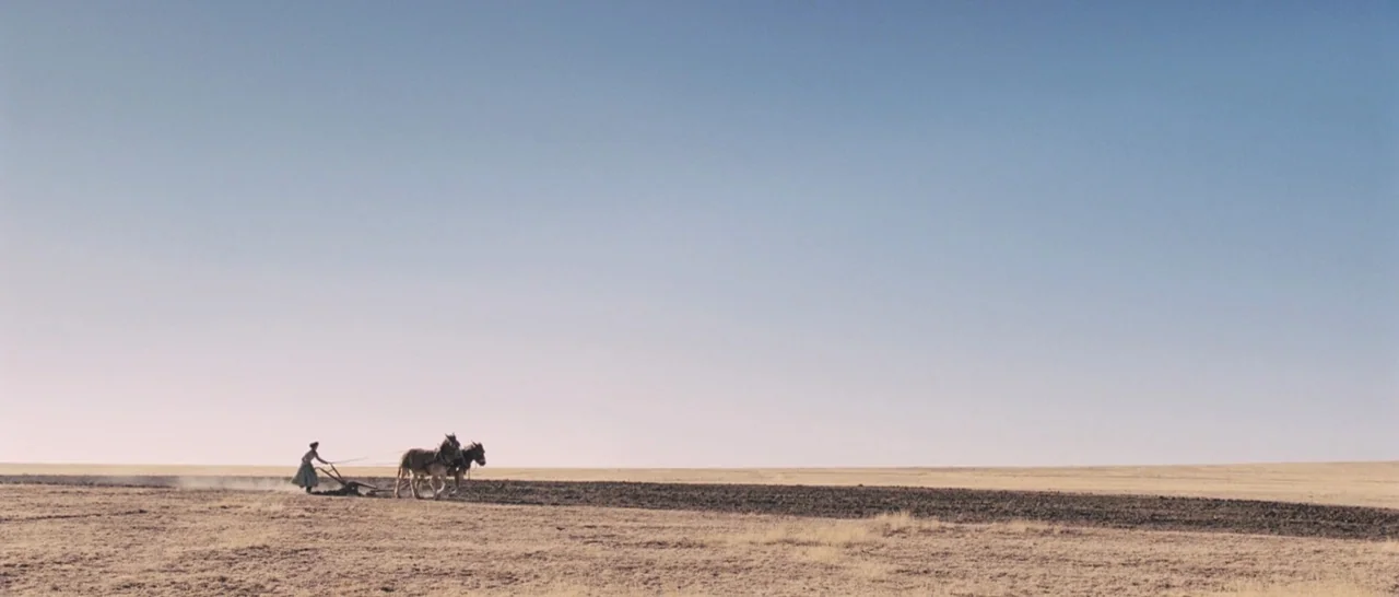 The Homesman (2014) movie still: wide — A vast, flat, arid field stretches toward the horizon under a clear sky. A lone woman uses a horse-drawn…