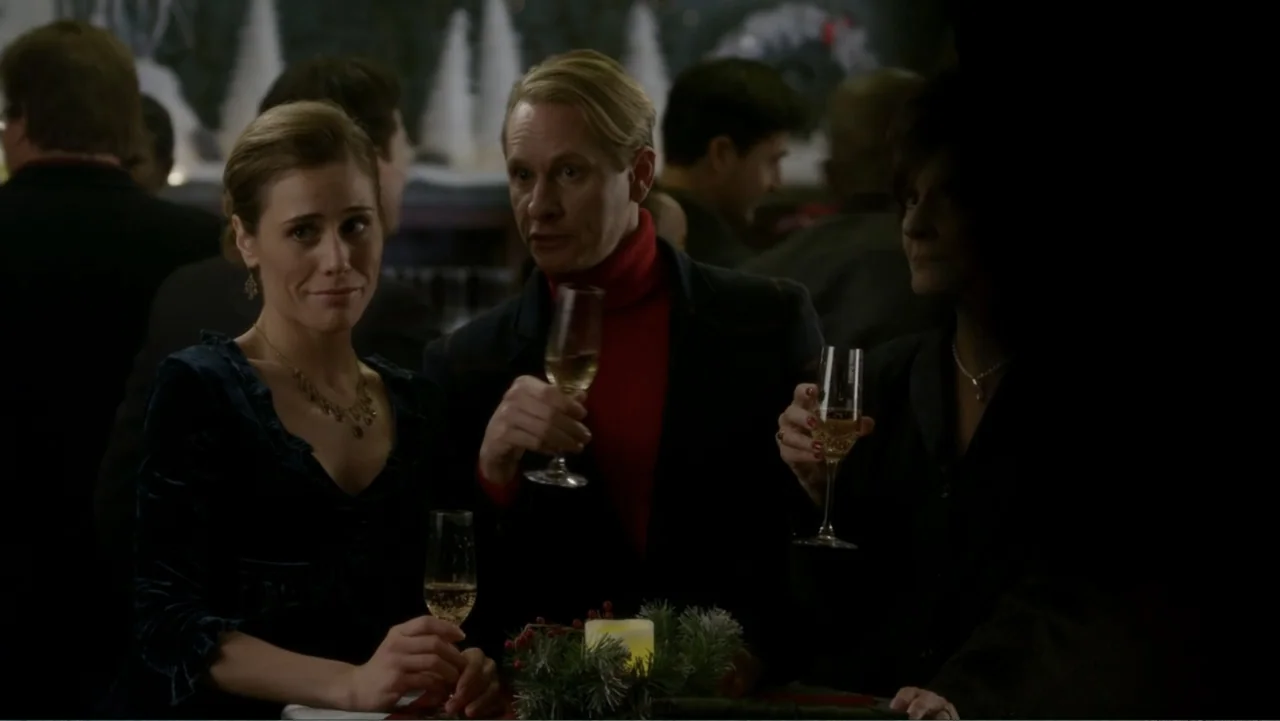It's Christmas, Carol! (2012) movie still: medium — A social gathering takes place in a dimly lit room with holiday decorations. Three people stand…