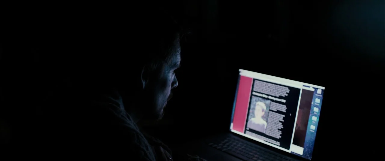 Sinister (2012) movie still: medium — A dark, dimly lit environment features a person viewing a screen in shadow. The subject looks at an article…