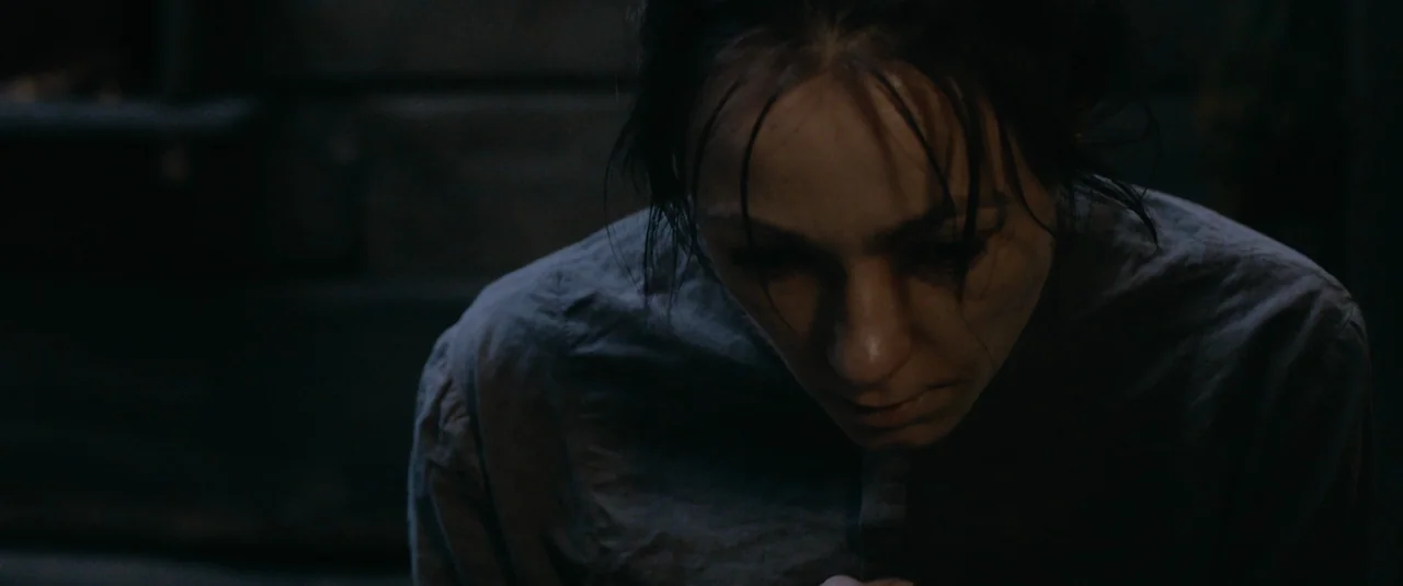 Hagazussa (2017) movie still: closeup — A dark, dimly lit interior space with a muted color palette. A woman with dark hair looks downward in a…