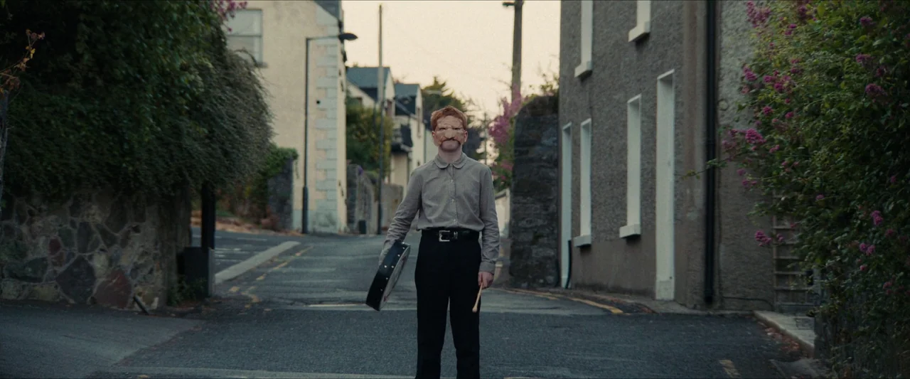 Fréwaka (2024) movie still: fullbody — A quiet residential street lined with old stone buildings and flowering bushes at dusk. A young man wearing a…