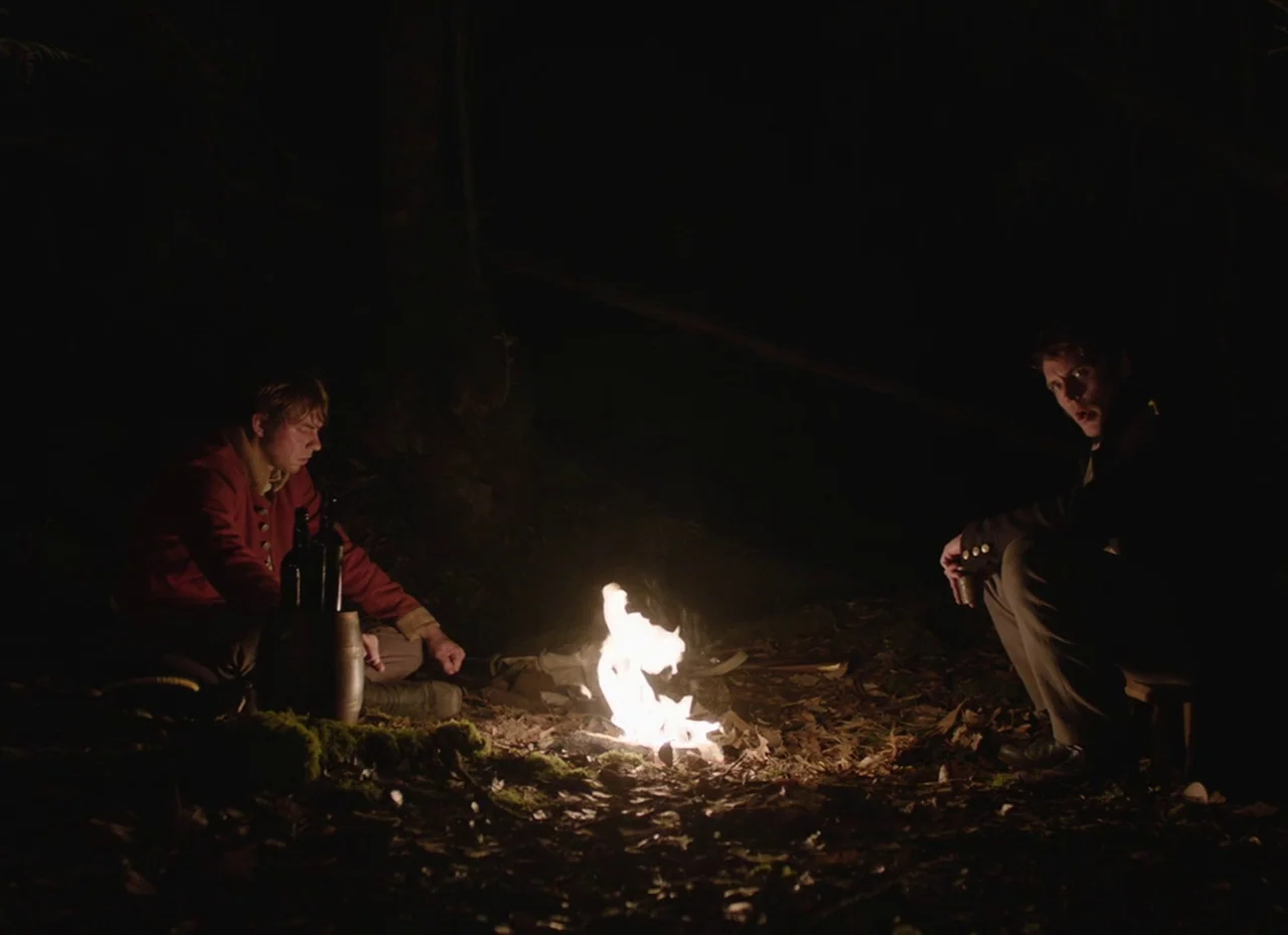 The Nightingale (2018) movie still: wide — A dark, wooded clearing at night with fallen leaves on the ground. Two men in historical uniforms sit…