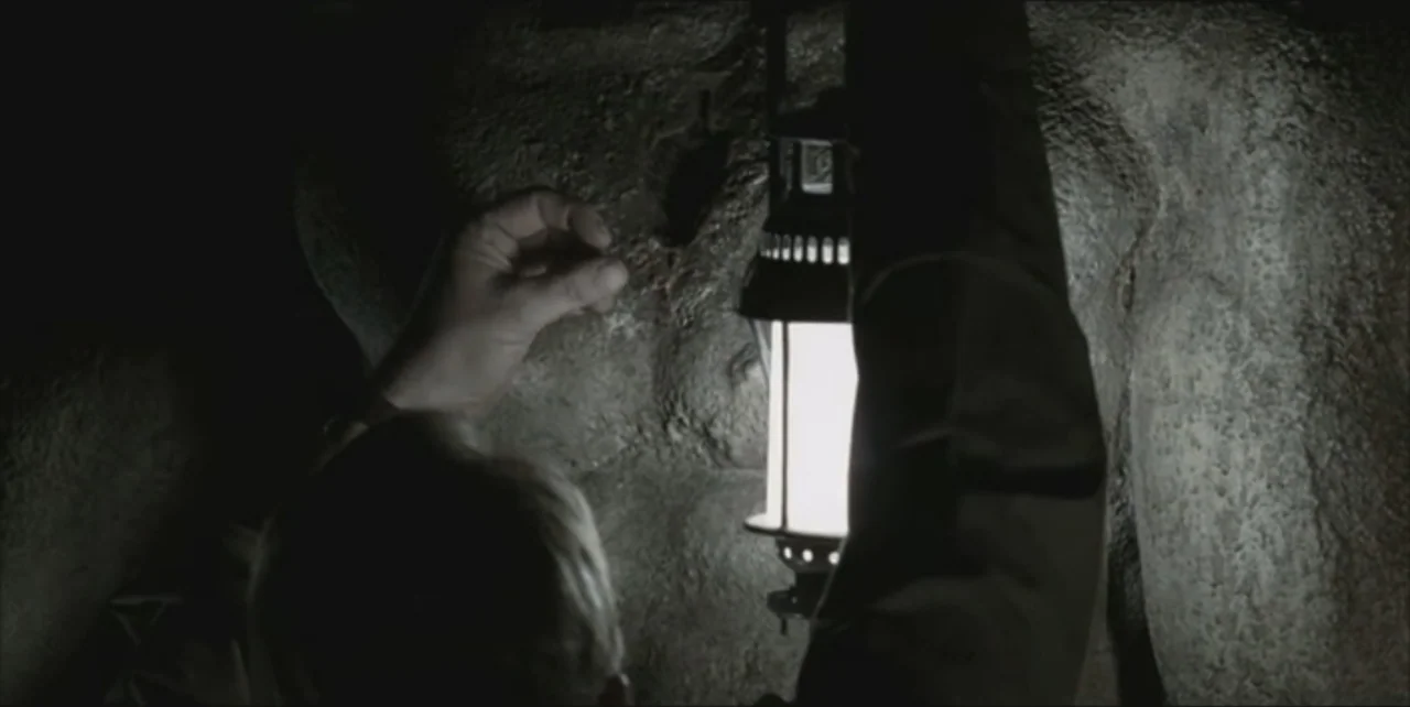 Exorcist: The Beginning (2004) movie still: closeup — A dark rocky cave interior features damp, uneven stone walls illuminated by a lantern. An…