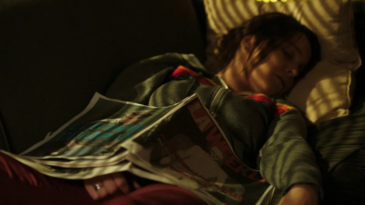 The Pact II movie still: medium — A person rests on a dark couch under the shadow of venetian blinds. A newspaper lies across their midsection while…