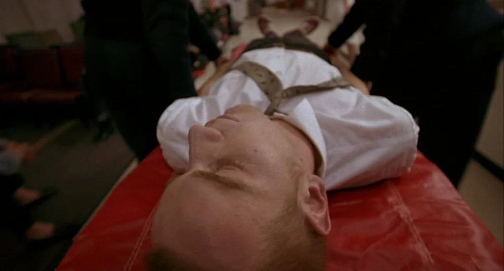 Trainspotting (1996) movie still: medium — A man lies on a red medical stretcher in a brightly lit corridor. The camera is positioned directly above…