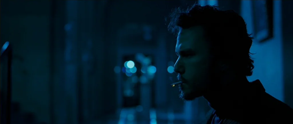 I’m Not There (2007) movie still: closeup — A dark, blurred corridor is illuminated by a single faint, cool blue light. A man with curly hair and a…