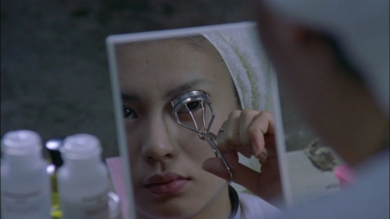 Battle Royale (2000) movie still: closeup — A close-up view shows a young woman reflected in a small handheld vanity mirror. She uses a metal…