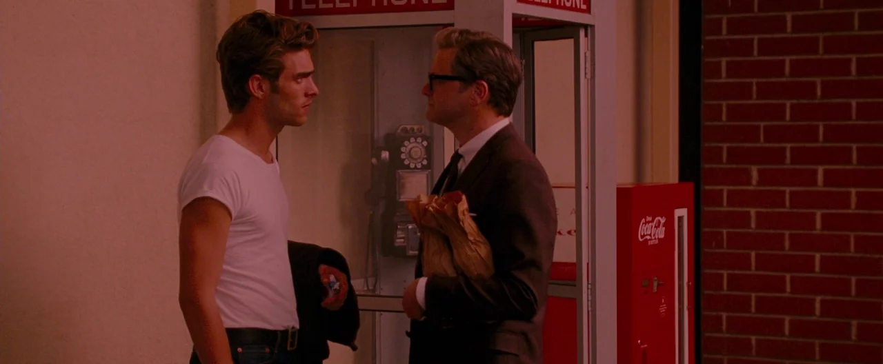 A Single Man (2009) movie still: medium — A man in a plain white t-shirt faces a middle-aged man in a suit. They stand beside a vintage telephone…
