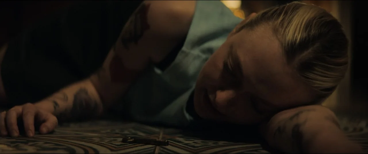 Vicious (2025) movie still: closeup — The interior setting features a patterned tiled floor with a single small metal key. A blonde woman lies prone…