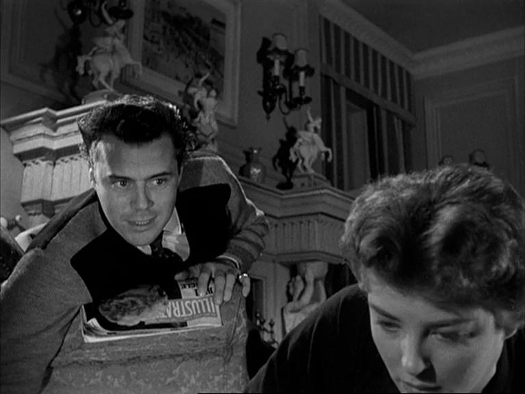 The Sleeping Tiger (1954) movie still: medium — A furnished living room features a fireplace mantle adorned with small white figurines. A man leans…