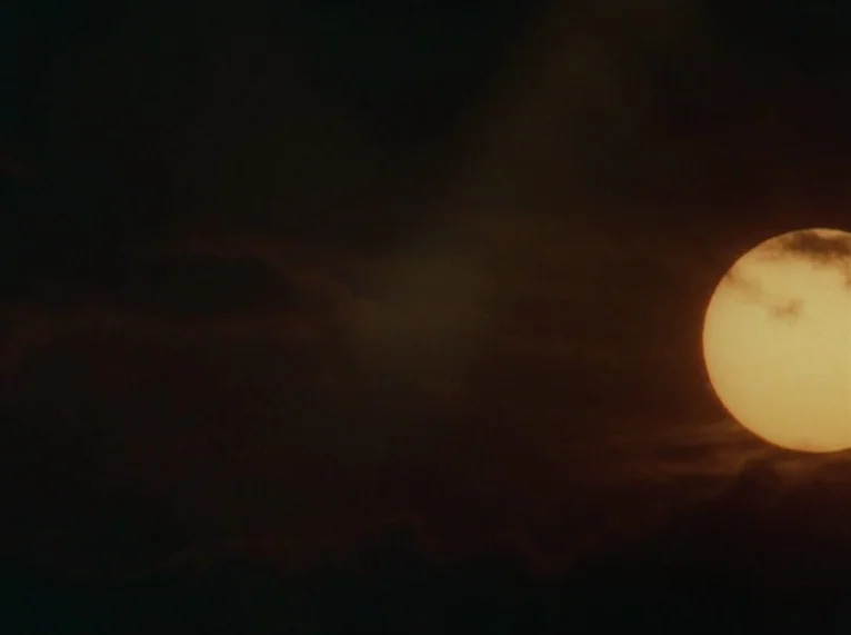 Hail Mary (1985) movie still: establishing — A dark, cloud-filled sky dominates the frame during a late sunset. The bright yellow sun is partially…
