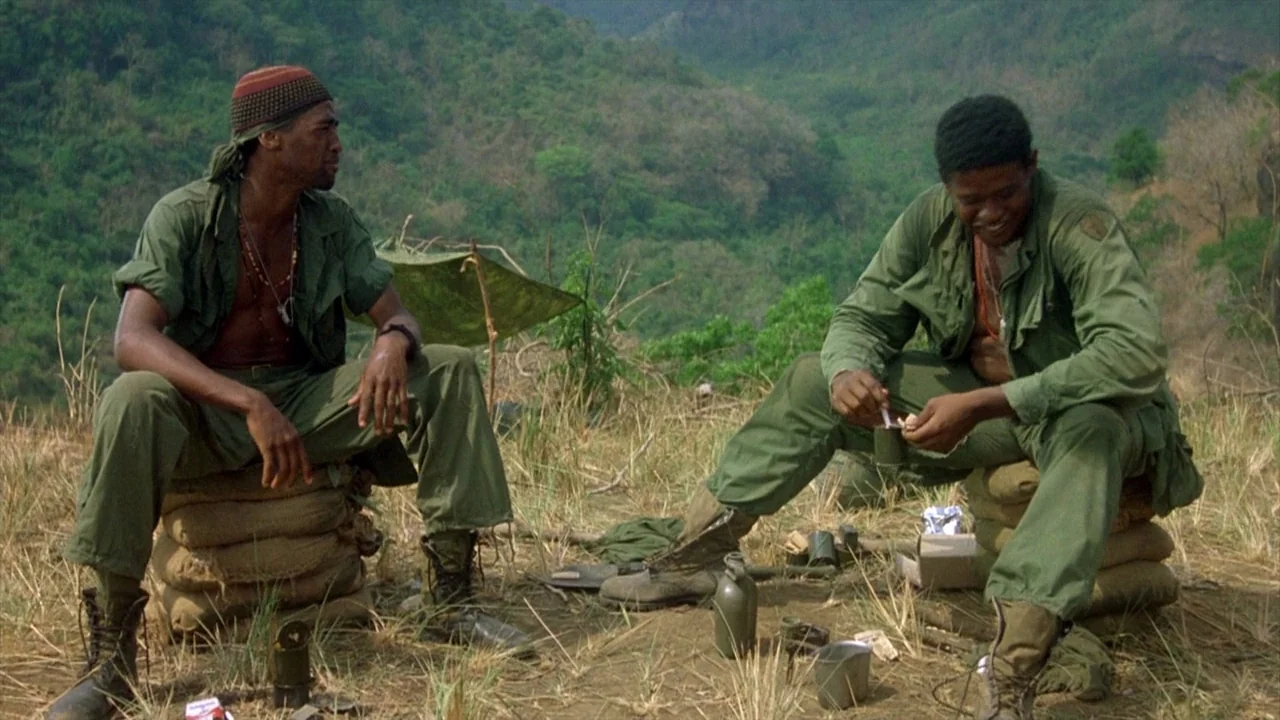 Platoon (1986) movie still: fullbody — A remote hillside outpost with dry grass and sandbag fortifications overlooking a distant mountain range. Two…
