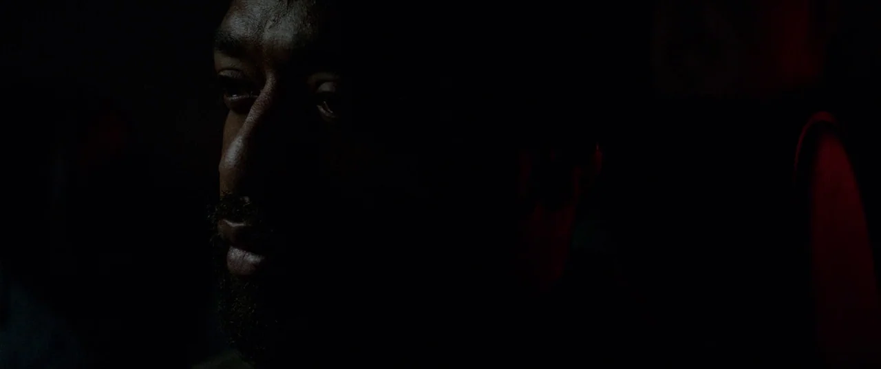 Triple 9 (2016) movie still: closeup — A dimly lit interior space with mostly obscured surroundings. A man with a short beard gazes into the dark…