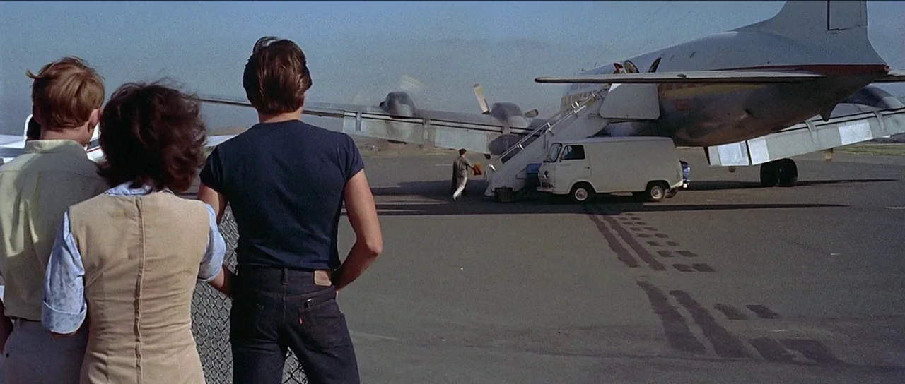 American Graffiti (1973) movie still: wide — A sunny airfield with a parked vintage twin-engine aircraft and a small service van. Three people stand…
