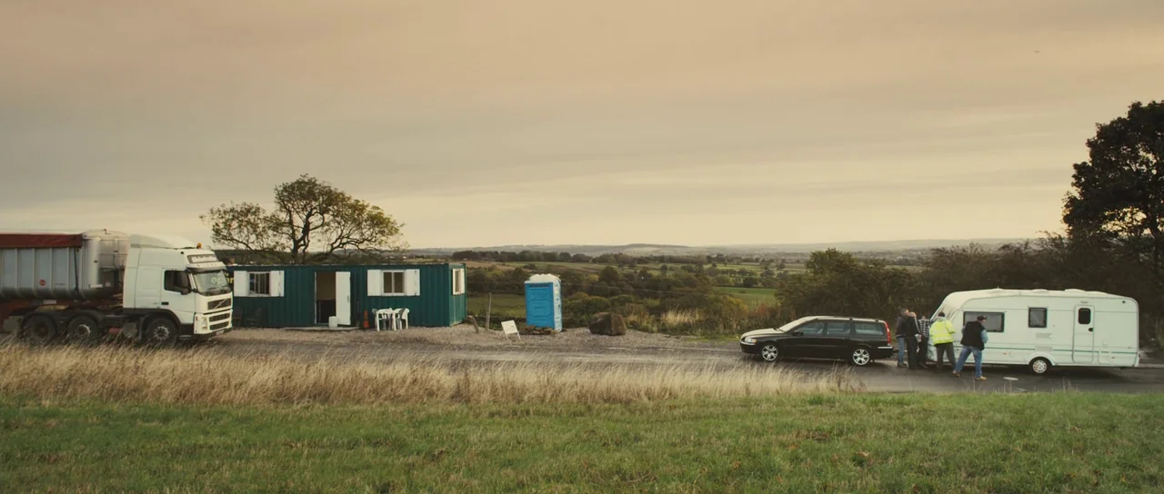 Sightseers (2012) movie still: wide — A rural roadside area features a parked truck, a temporary office container, and a caravan. A small group of…