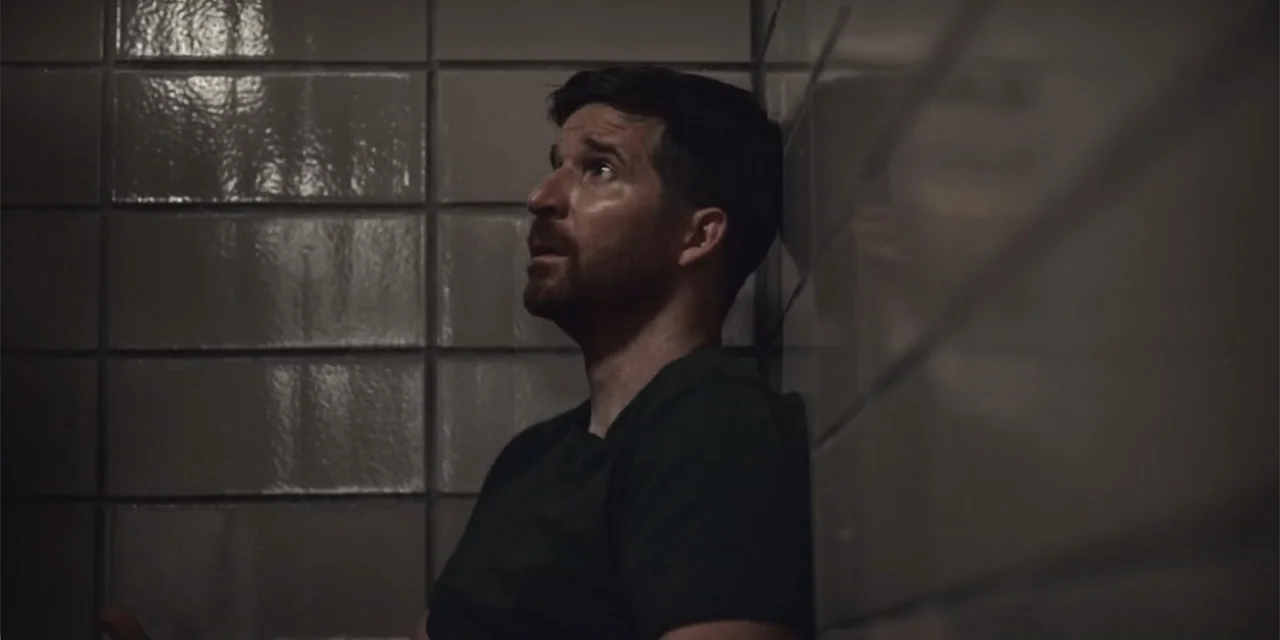 Equal Standard (2021) movie still: medium — A corner of a room featuring large, glossy gray tiles on the walls. A man in a dark shirt rests against…