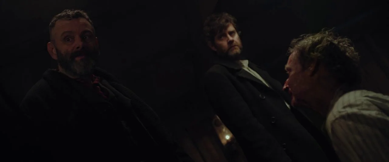 Apostle (2018) movie still: medium — A dimly lit interior space with rough wooden walls and shadows. Three men gather in a dark room looking down at…