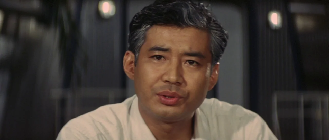 Son of Godzilla (1967) movie still: closeup — An interior space with dark, panelled walls featuring recessed wall sconces. A man in a light shirt…