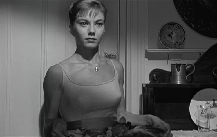 On The Beach (1959) movie still: medium — A woman stands in a dining room with a white door and wood furniture. She holds a large platter of food…