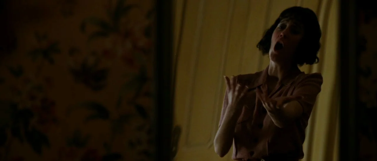 La Vie En Rose (2007) movie still: medium — A dimly lit residential hallway features dark floral wallpaper and a paneled wooden door. A woman with…