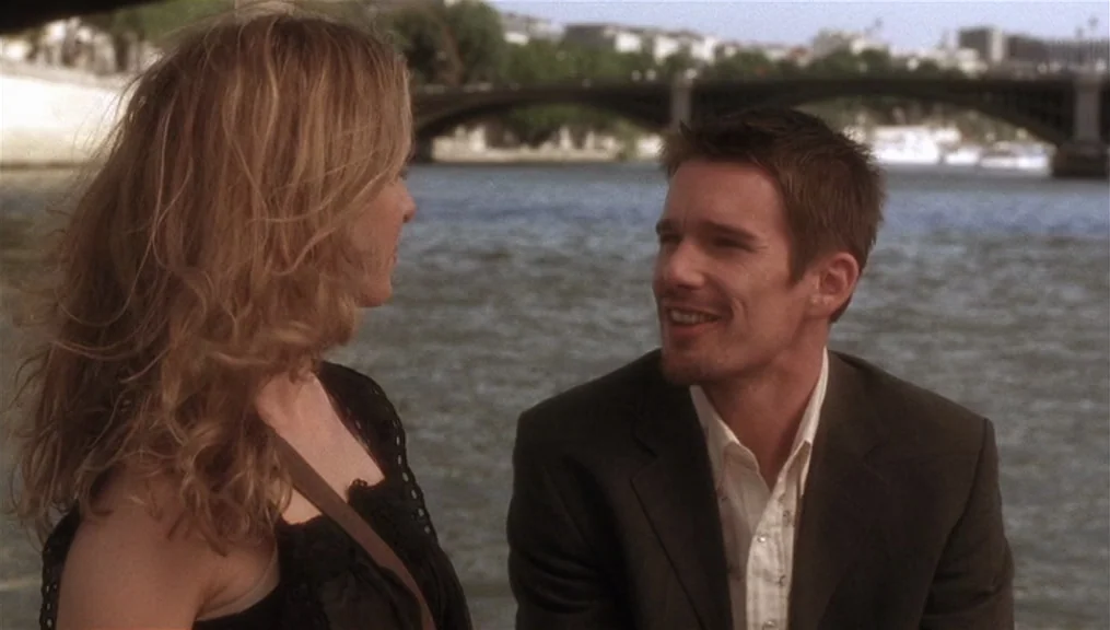 Before Sunset (2004) movie still: medium — A man and woman sit along a riverbank with a stone bridge in the distance. The pair faces each other…