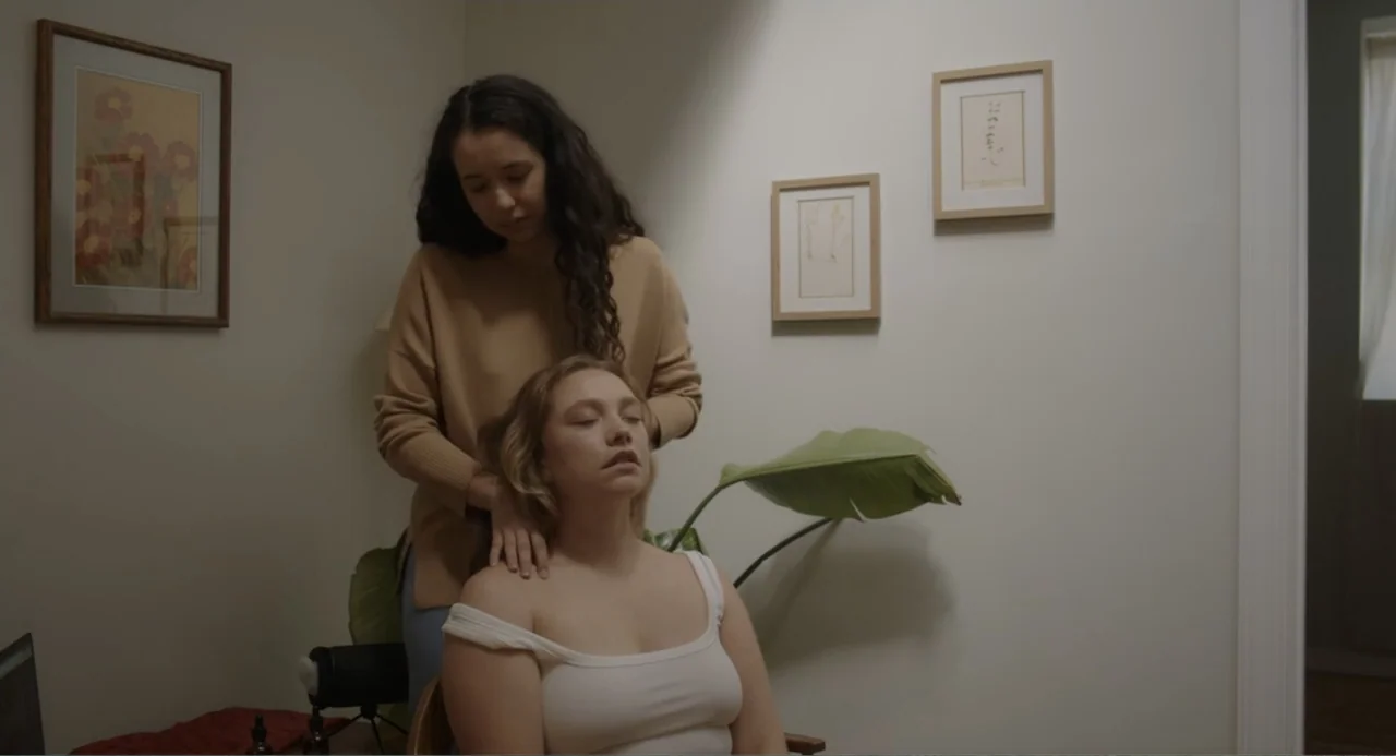 This Closeness (2024) movie still: medium — An interior space with off-white walls decorated with framed botanical art prints and a plant. One woman…