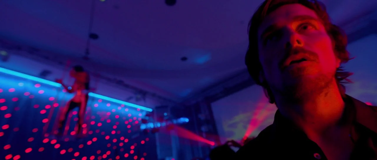 Knight of Cups (2015) movie still: closeup — A dark, neon-lit room features blurred red and blue light patterns across the space. A man gazes upward…