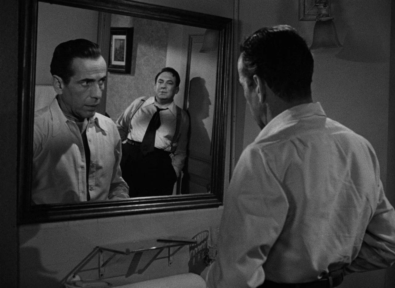 Key Largo (1948) movie still: medium — A small room features a large rectangular mirror hanging above a tiled basin area. Two men in shirts observe…