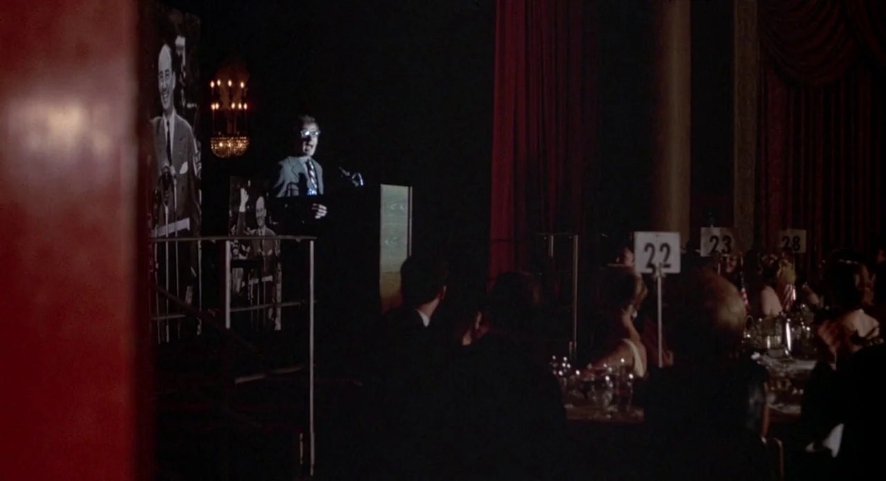 Annie Hall (1977) movie still: wide — A formal banquet hall featuring red velvet curtains and round dinner tables. A man in a suit speaks into a…