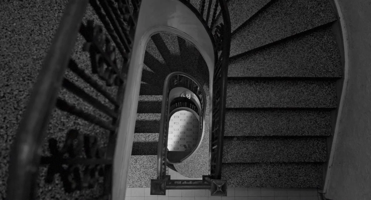 Song of Granite (2017) movie still: establishing — A stone spiral stairwell with dark metal railings descends deep into the building core. The view…