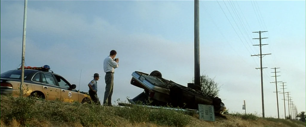 Fight Club (1999) movie still: wide — A rural roadside scene features an overturned sedan lying against a wooden utility pole. A man in a dress…