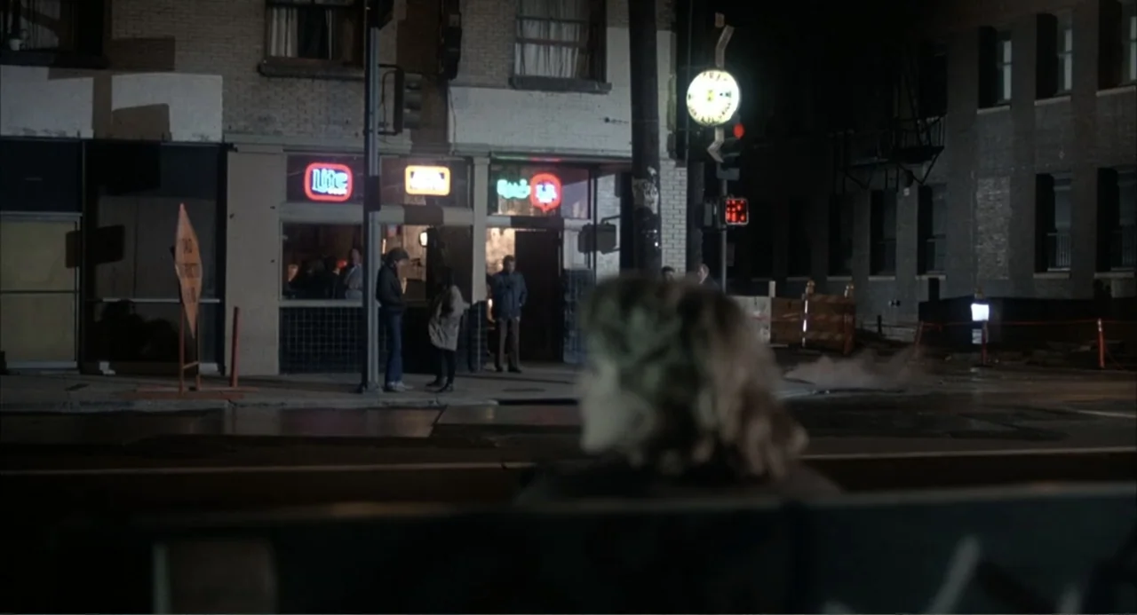 I, Madman (1989) movie still: wide — A dark urban corner features a small commercial storefront with brightly glowing neon signage. Three people…
