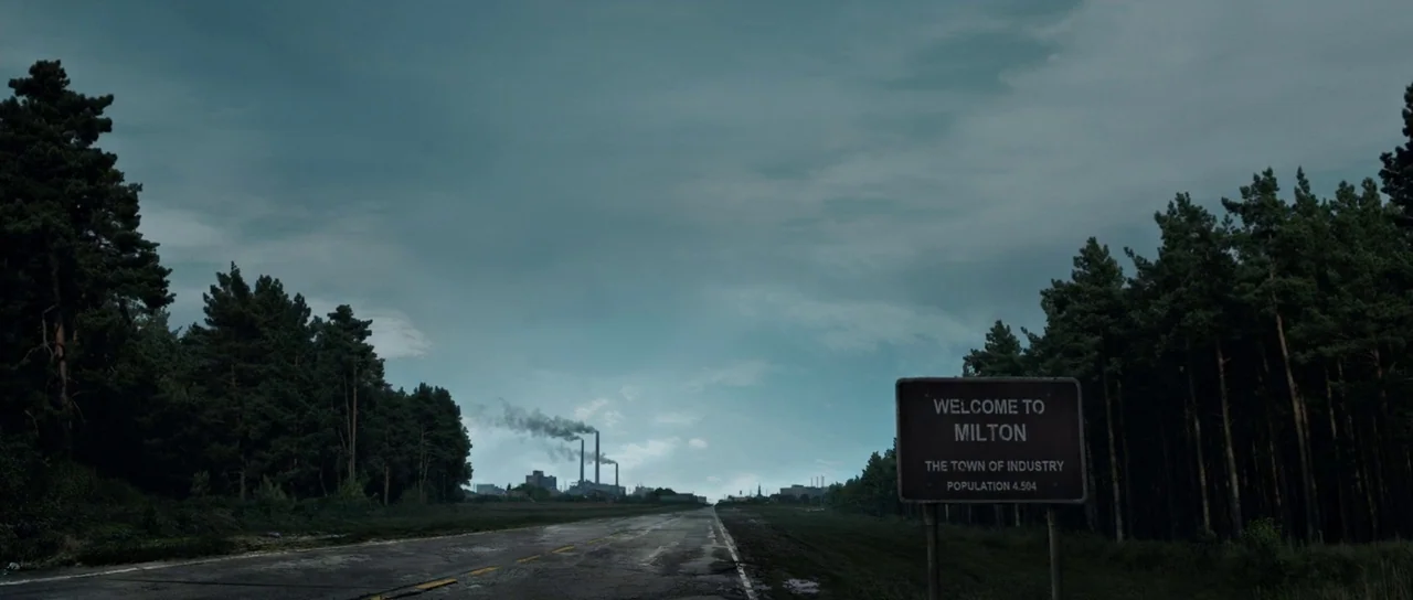 The Voices (2014) movie still: establishing — A desolate two-lane highway flanked by dense pine forests under an overcast sky. A rusted sign…