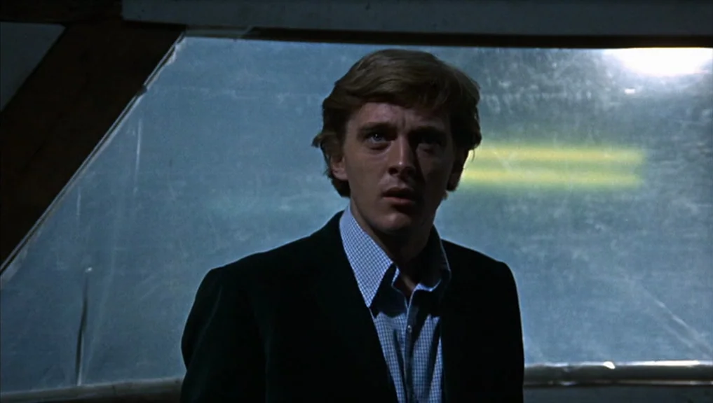 Blow-Up (1966) movie still: medium — A dimly lit interior space features a large, slanted rectangular window at the back. A young man stands in the…