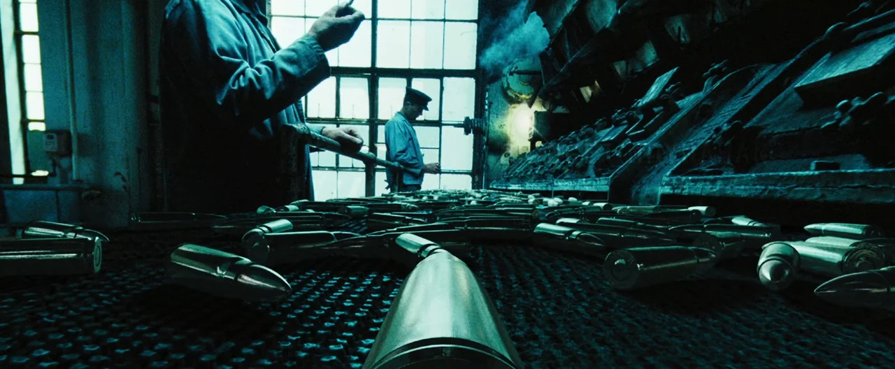 Lord of War (2005) movie still: medium — A dark industrial factory features a conveyor belt moving large metal bullets. Two male workers stand in…