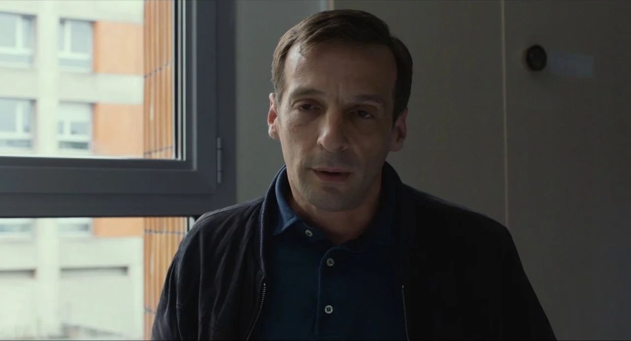 Happy End (2017) movie still: medium — A man stands in an office in front of a window overlooking a building. He is looking off-screen and appears…