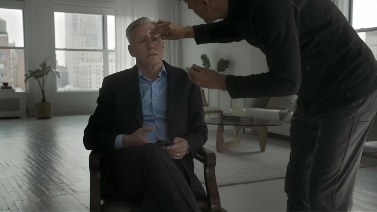 Predators (2010) movie still: medium — A minimalist living room with white walls and a large city window. A seated man has his forehead wiped with a…