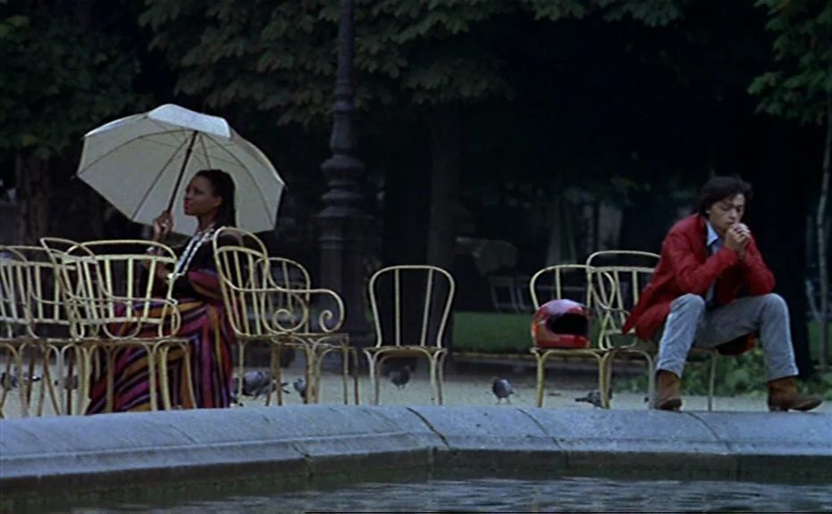 Diva (1981) movie still: wide — A public park featuring a circular stone fountain surrounded by rows of metal chairs. A woman with an umbrella and a…