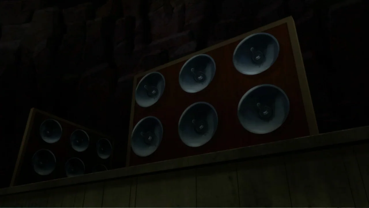Superman: Red Son (2020) movie still: medium — A dark, textured stone cave wall is partially obscured by two large speaker arrays. The speaker units…